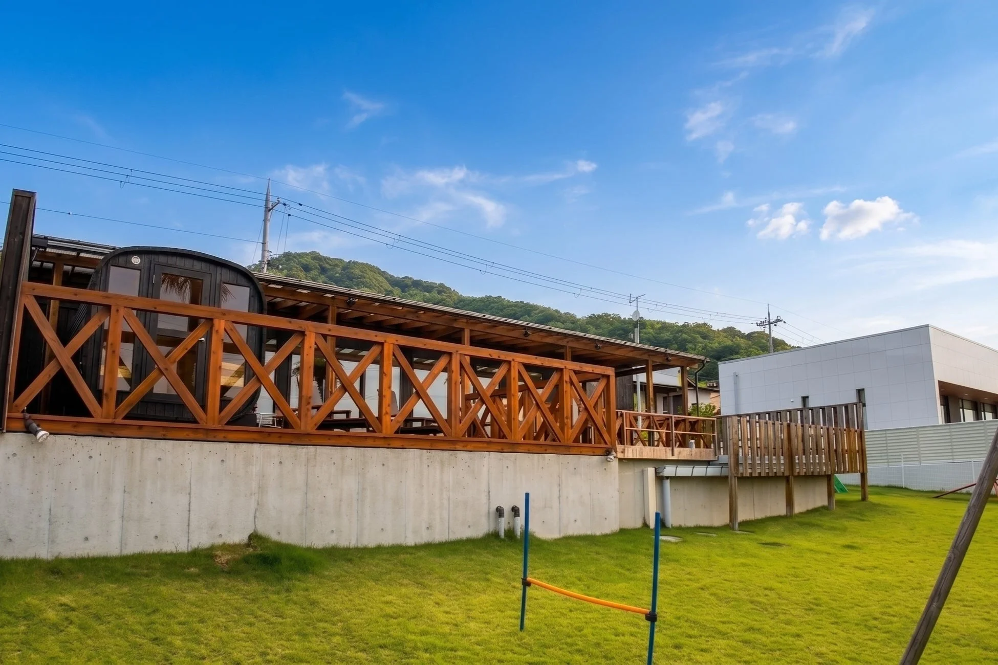 青い空と緑の芝生が広がる敷地に、木製の広いデッキがあるモダンな建物が建っています。デッキには特徴的な丸い形状の施設があり、リラックスできる空間が広がっています。