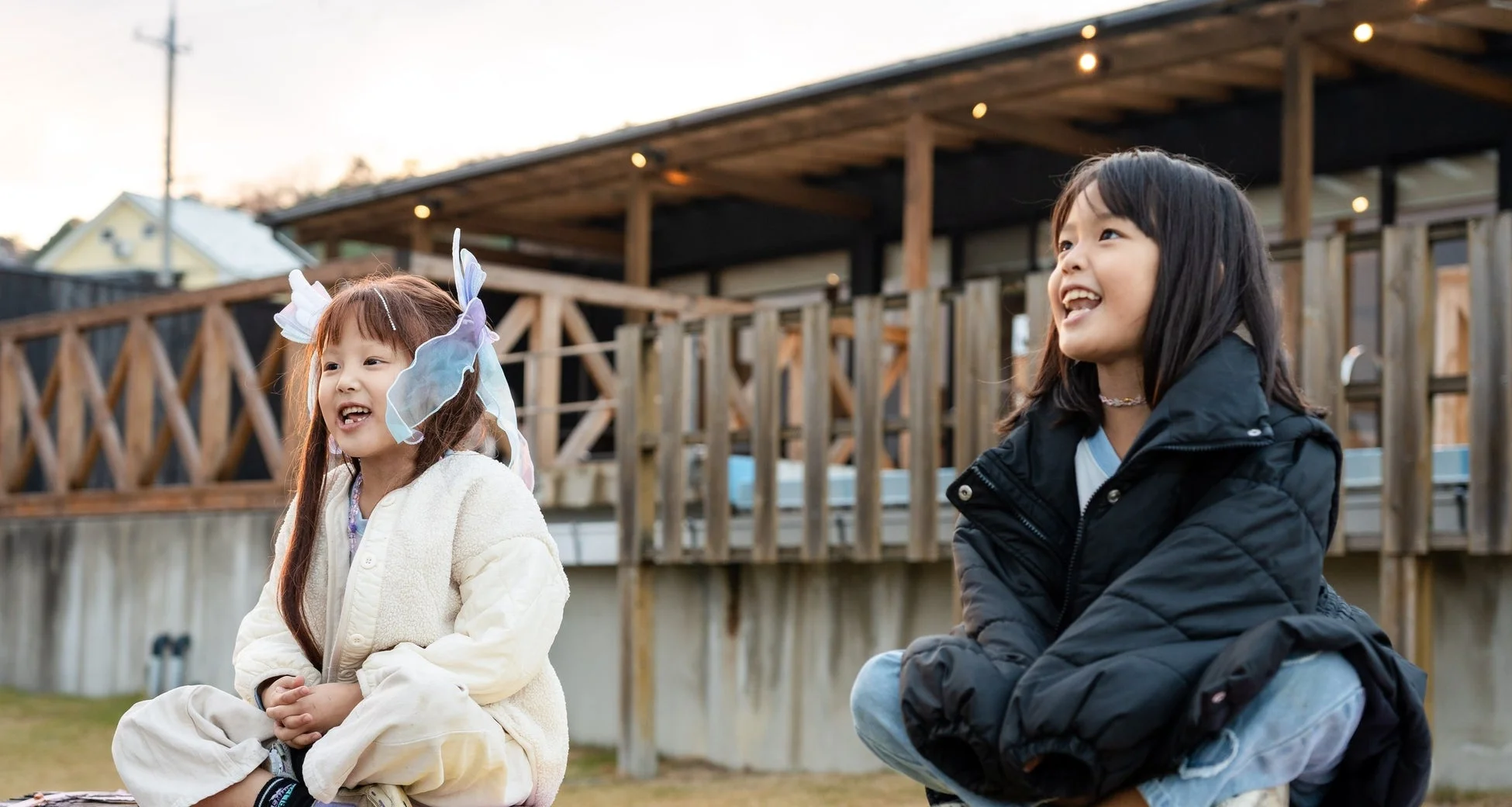 屋外で笑顔を見せる2人の幼い女の子が写っています。一人は黒いダウンジャケットを、もう一人は白いフリースジャケットと可愛らしい髪飾りを身につけ、楽しそうに座っています。背景には木製のフェンスと建物が見えます。
