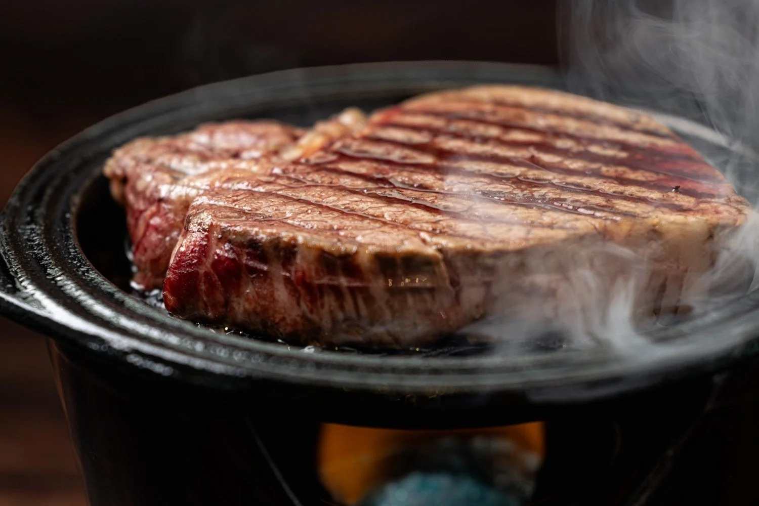 熱々の鉄板の上で、美しい焼き目がついた厚切りのステーキが湯気を立てながら焼かれている様子です。ジューシーな肉質が食欲をそそります。