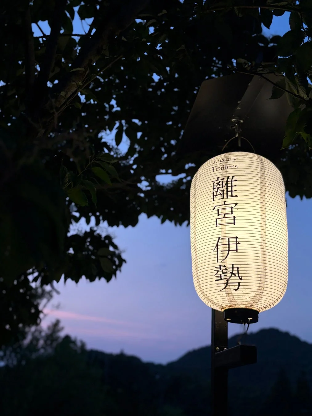 夕暮れ時、暗がりの木々と山々を背景に、「離宮伊勢」と書かれた和風の提灯が明るく灯されている。