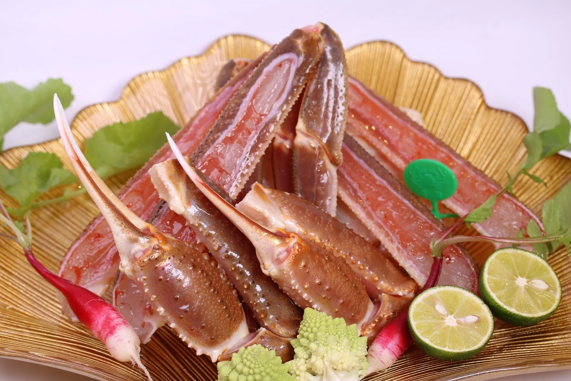 金色のお皿に盛り付けられた新鮮なカニの脚肉の刺身です。身が詰まったカニの脚が美しく並べられ、彩りとしてライム、ラディッシュ、緑の葉物野菜が添えられています。高級感のある海鮮料理の盛り付けです。