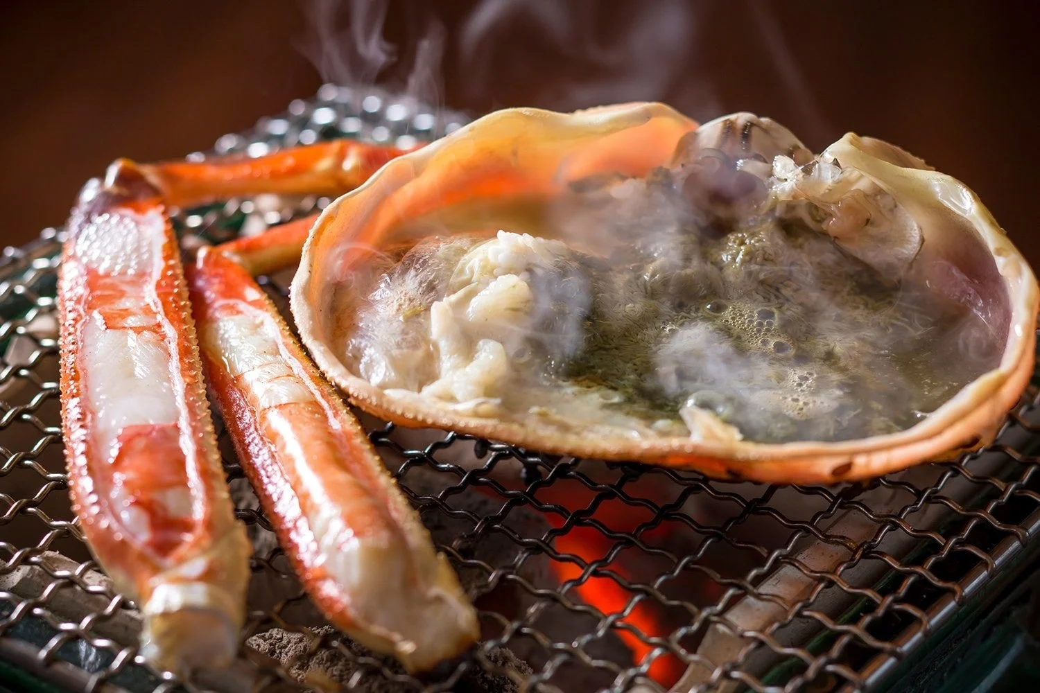 網の上で熱々に焼かれるカニの脚と甲羅焼き。湯気が立ち上り、香ばしい香りが食欲をそそる、冬の味覚を代表する贅沢な一品です。