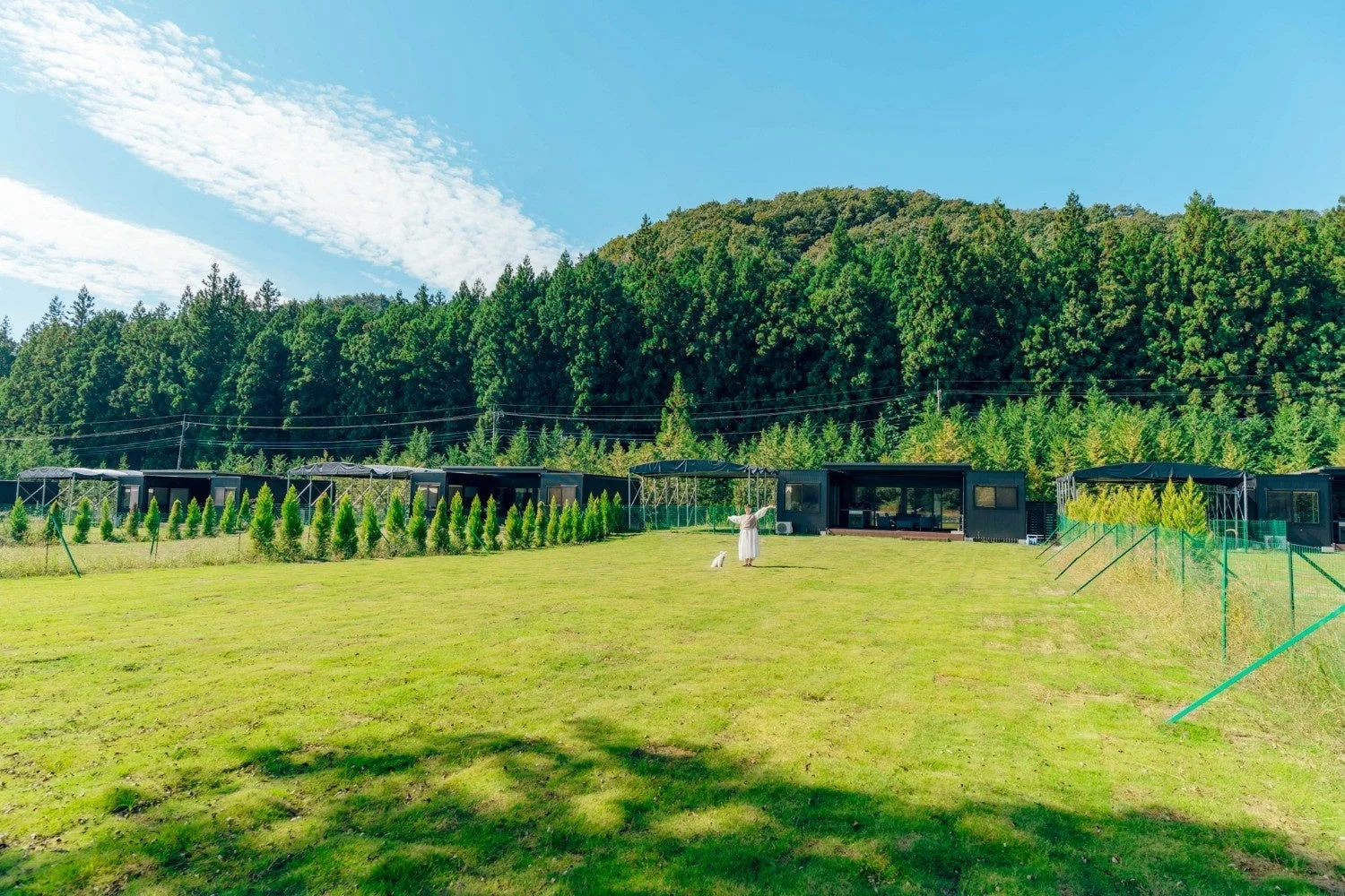 広々とした芝生が広がる敷地にモダンな宿泊施設が並び、一人の女性と白い犬がくつろいでいます。背景には豊かな緑の森と青い空が広がる、自然に囲まれた穏やかな風景です。