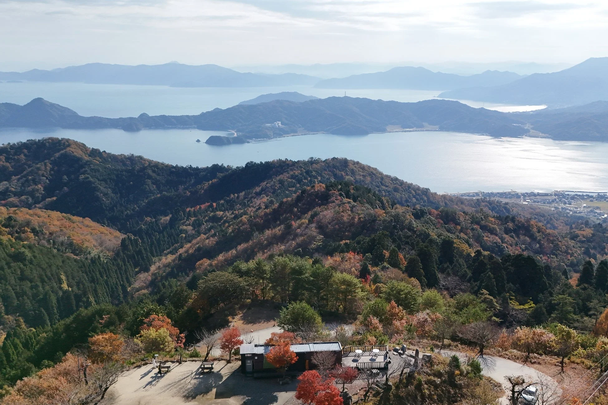 秋の紅葉に彩られた山々が広がる絶景で、手前には展望台らしき建物が見えます。遠くには穏やかな海に浮かぶ島々が広がり、澄み渡った空の下、壮大な自然のパノラマが広がっています。