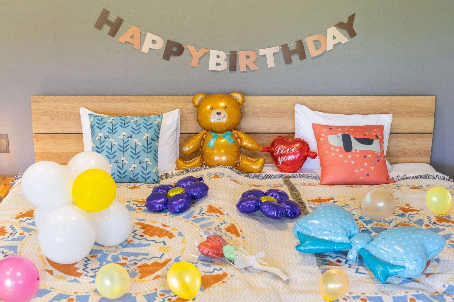 ベッドの上に誕生日を祝うための飾り付けがされた写真です。HAPPY BIRTHDAYのバナーや、テディベア、ハート、様々な色のバルーンが並べられ、華やかな雰囲気を演出しています。