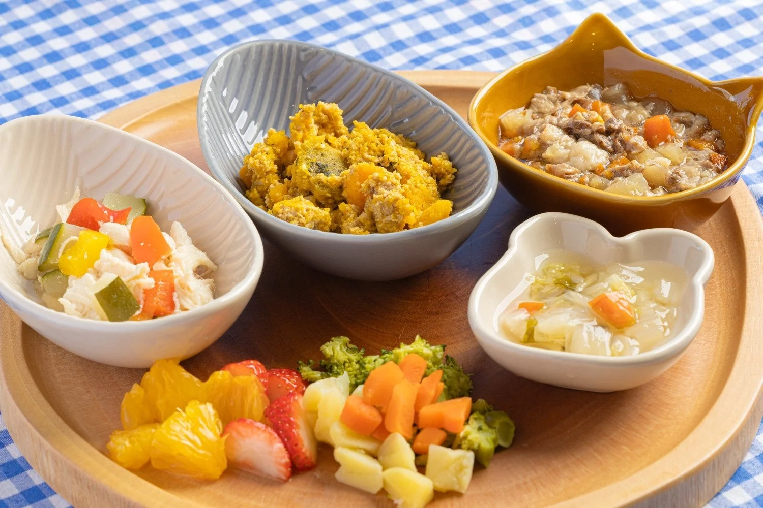 木製トレイに、複数の小鉢に入った栄養満点なペット用手作り食が並んでいます。肉、野菜、フルーツがバランス良く盛り付けられ、見た目も鮮やかで健康的な食事が青いチェックのテーブルクロスの上で提供されています。