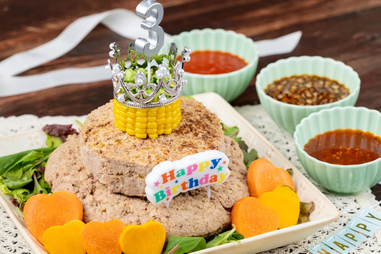 3歳の誕生日を祝うための、ミートローフのような手作りケーキが写っています。小さな王冠と「Happy Birthday」の飾り付けがあり、ハート形にカットされた野菜や数種類のソースが添えられています。